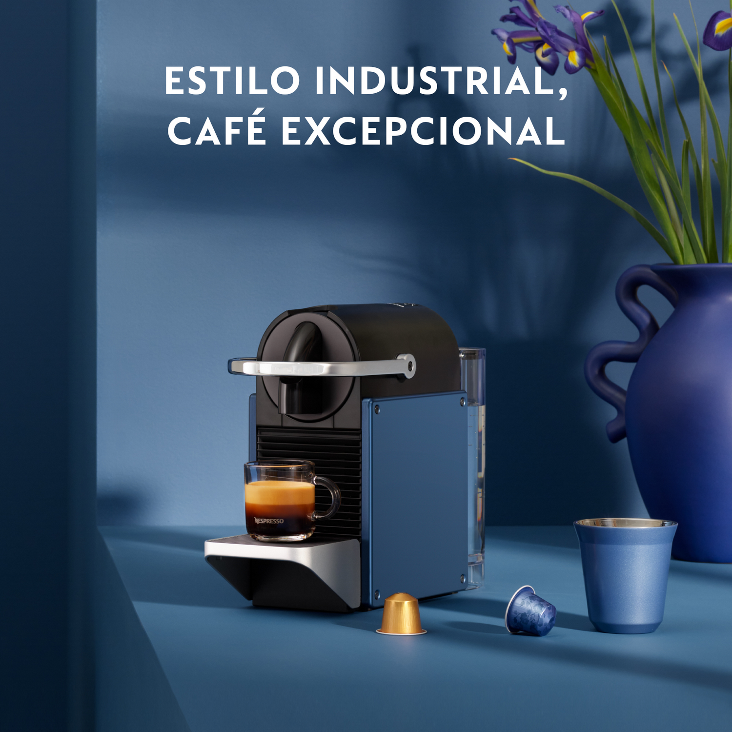 Una cafetera azul y negra con un vaso de café, cápsulas de café, una taza azul y un jarrón de flores sobre una superficie azul.