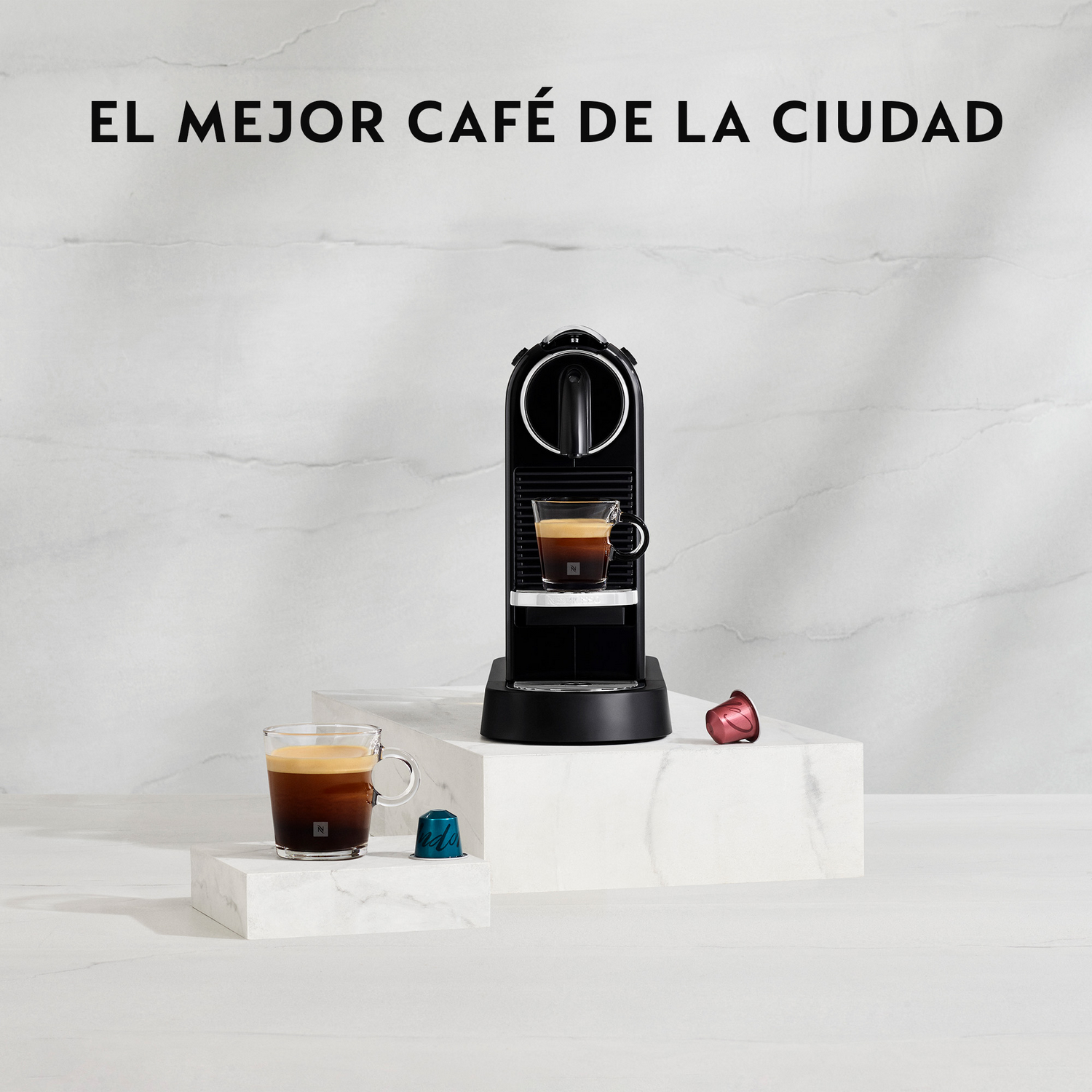 Máquina de café negra con dos tazas de espresso. Dos cápsulas de café están al lado de la máquina. El fondo es blanco con textura de mármol.