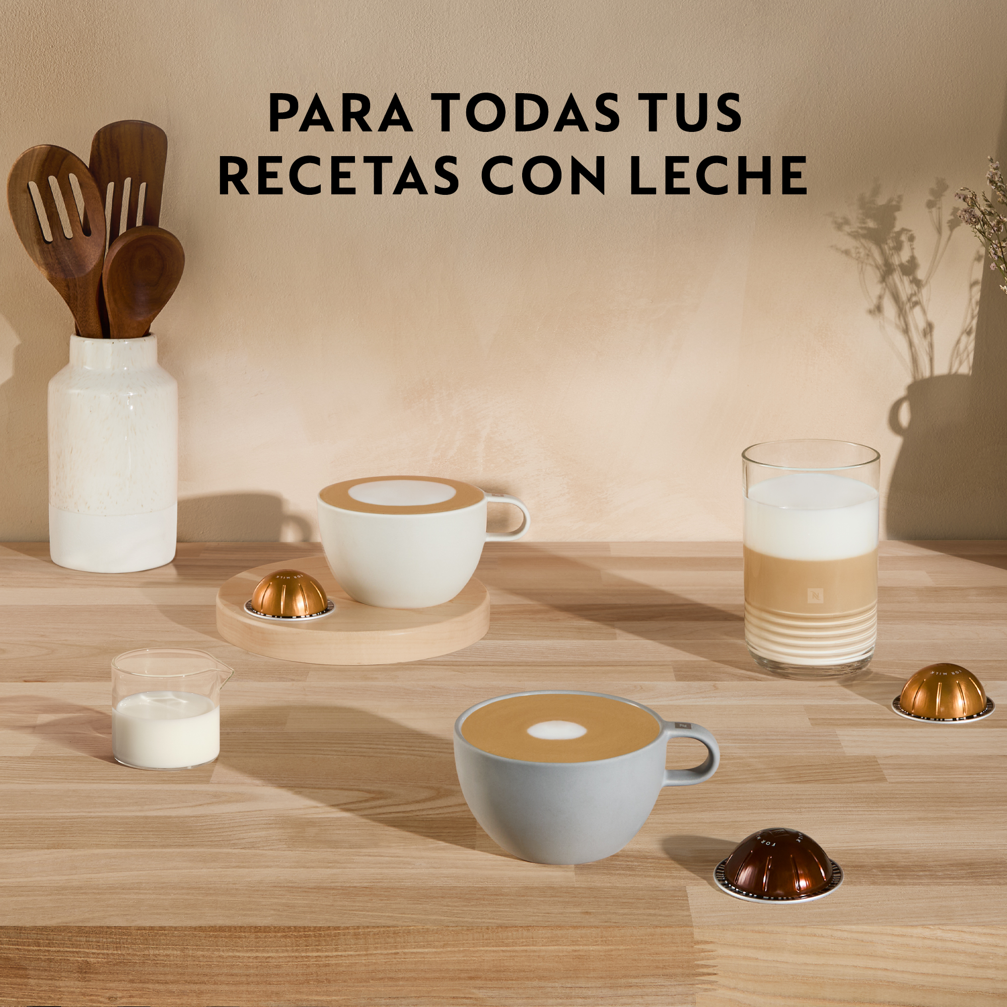 Tazas de café, cápsulas y leche sobre una mesa de madera, con texto 'PARA TODAS TUS RECETAS CON LECHE'.