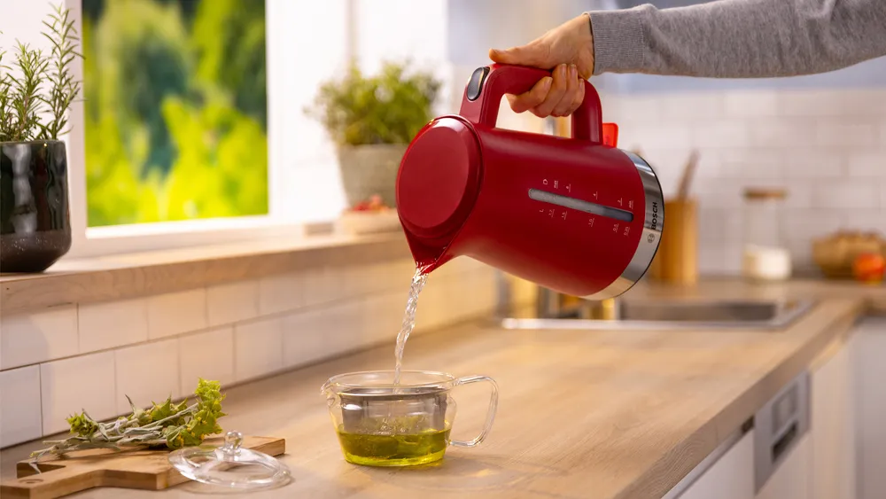 Egy piros vízforraló vizet önt egy üveg teáscsészébe, amiben tea van. Fűszernövények és növények egy fa pulton, ablak mellett.