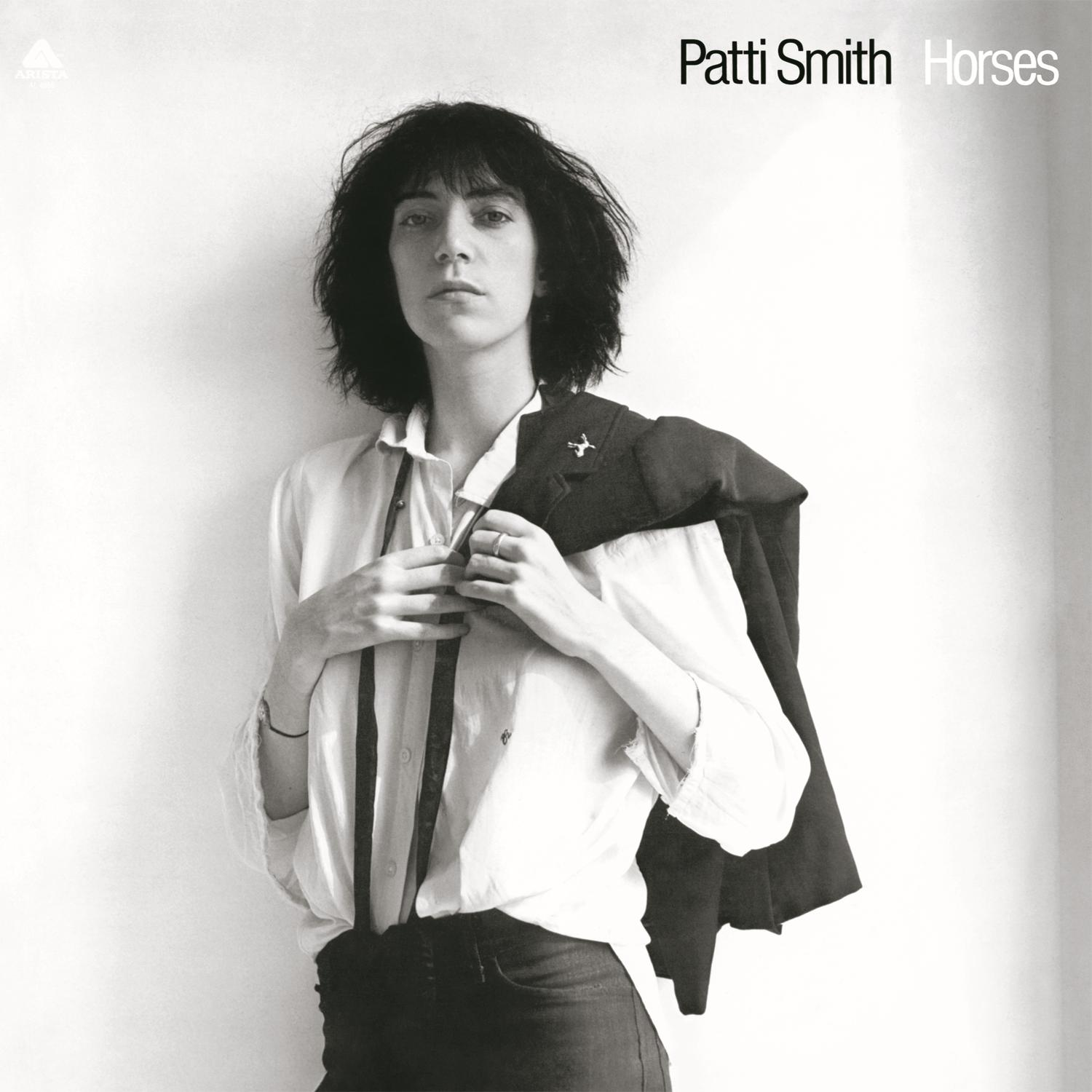 Fekete-fehér fotó Patti Smith-ről. Fehér inget, sötét nadrágot és a vállán egy kabátot visel, a nézőre néz.