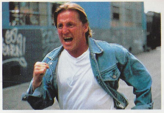 Ein Mann in einer Jeansjacke und einem weißen T-Shirt rennt mit aufgeregtem Gesichtsausdruck. Graffiti ist auf einer Wand im Hintergrund zu sehen.