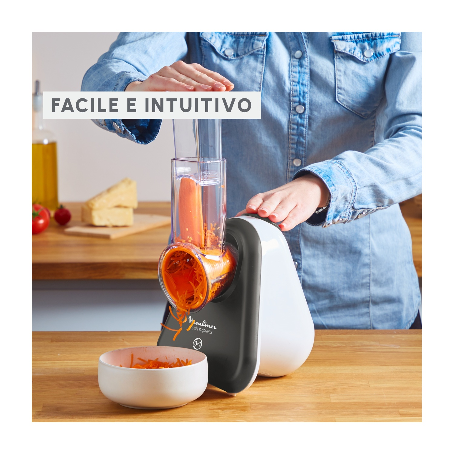 Una persona usa un Moulinex Fresh Express per grattugiare una carota. La carota grattugiata cade in una ciotola bianca. Sfondo: formaggio, pomodori, olio.