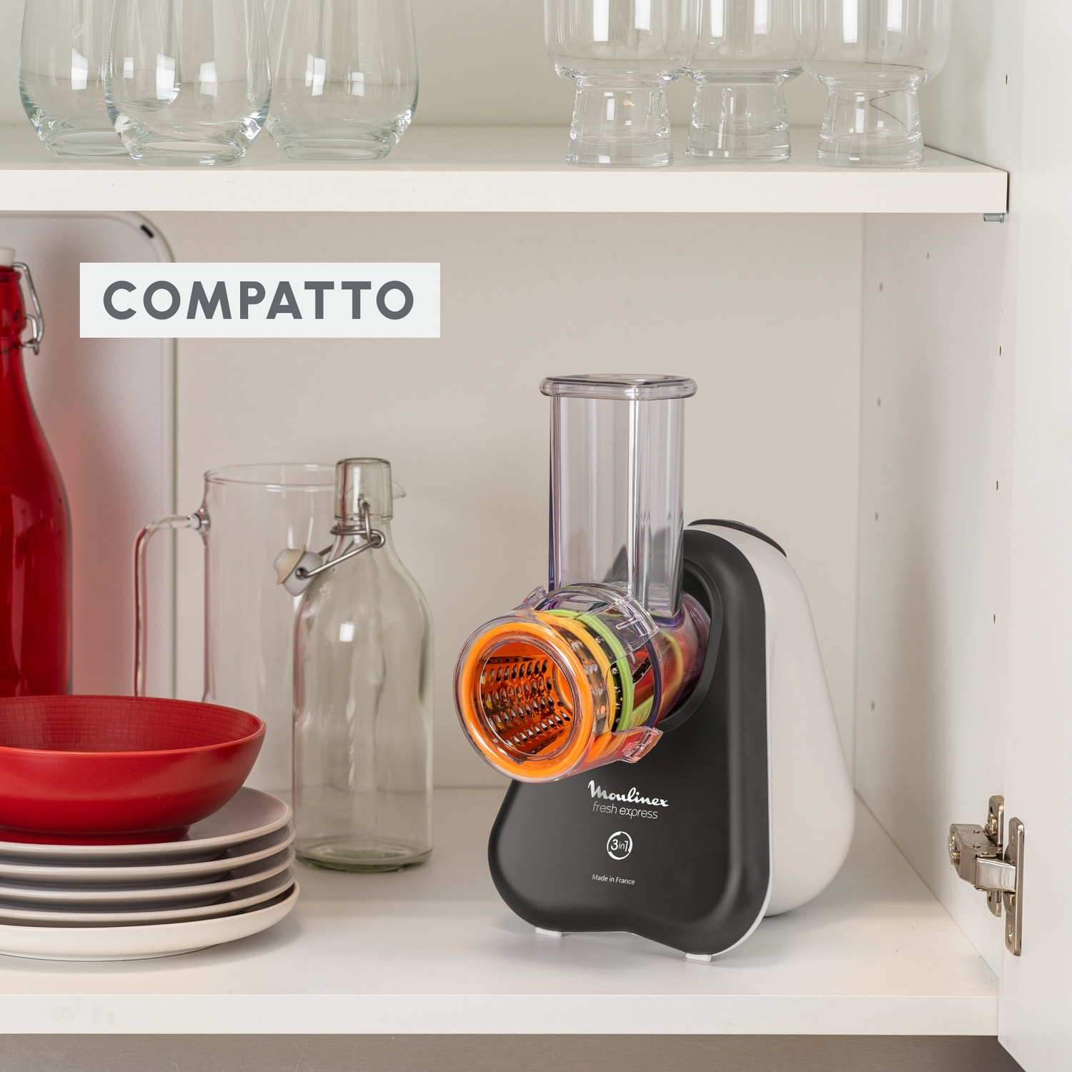 Un robot da cucina Moulinex Fresh Express si trova all'interno di un armadio. Una ciotola e una bottiglia rosse sono a sinistra. Sopra vetreria trasparente.