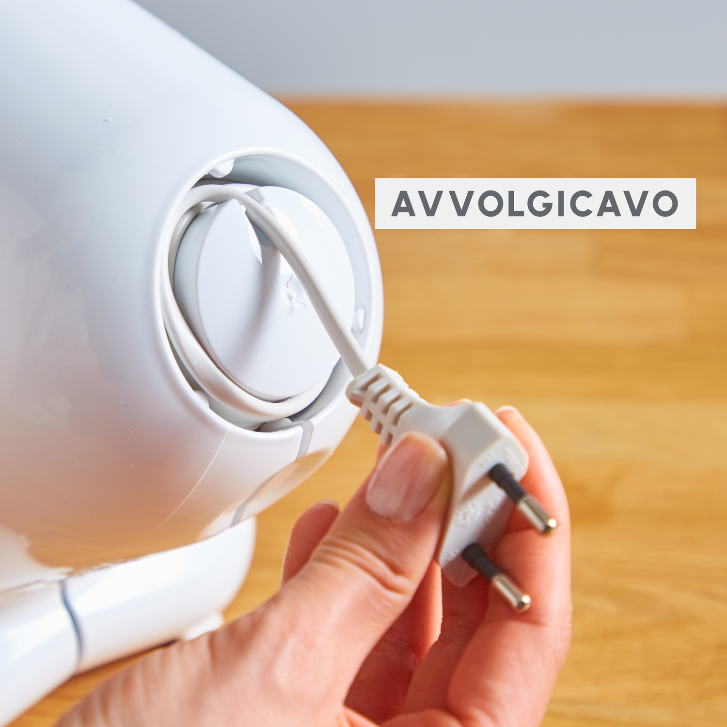 Una mano tiene una spina bianca e la collega a un dispositivo bianco. Il dispositivo ha un cavo all'interno, lo sfondo è in legno.