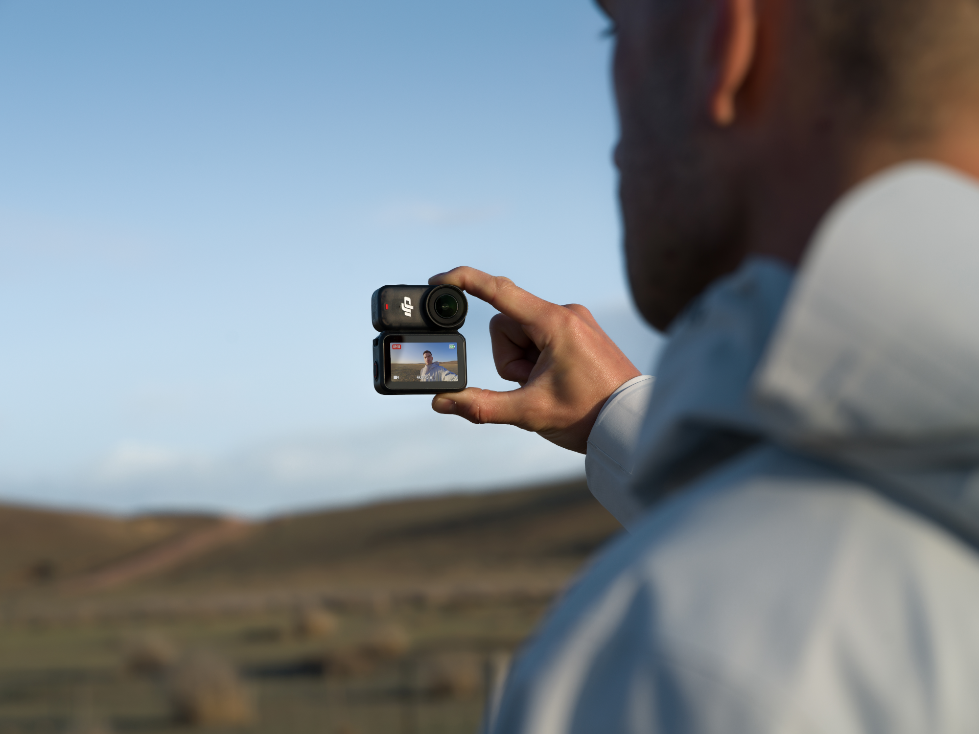 Eine Person hält eine Kamera und nimmt eine Landschaft unter blauem Himmel auf. Die Kamera zeigt eine Live-Ansicht der Person.