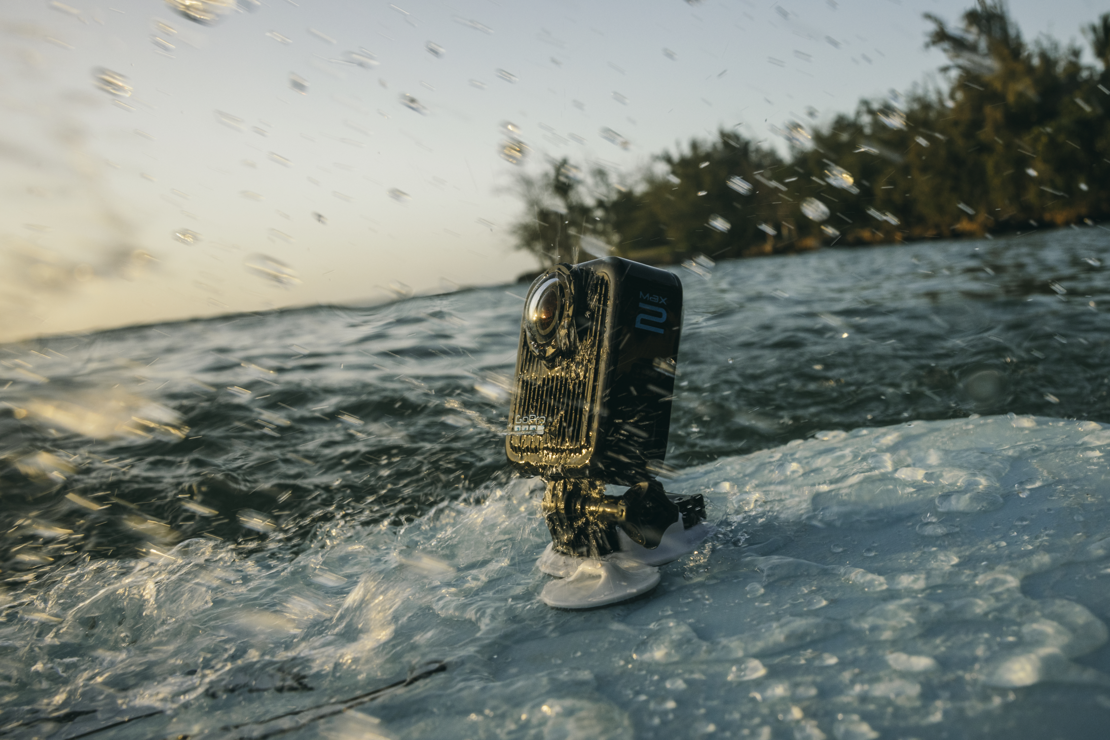 Eine schwarze GoPro-Kamera auf einer Halterung im Wasser mit Spritzern. Im Hintergrund Wasser, Bäume und ein heller Himmel.