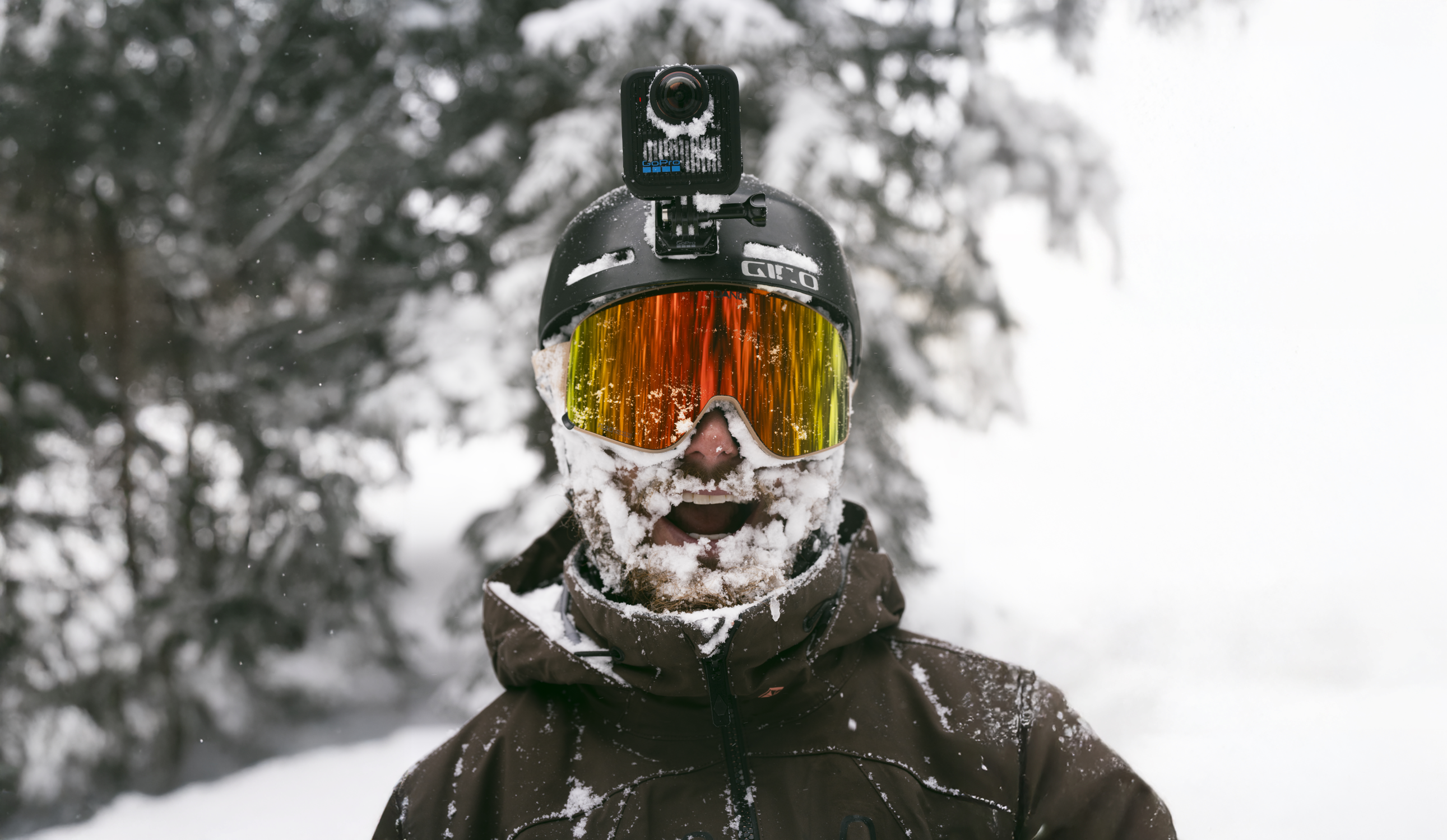 Eine Person in einer verschneiten Umgebung, die einen Helm mit Kamera, Skibrille und Jacke trägt. Schnee bedeckt Gesicht und Bart.