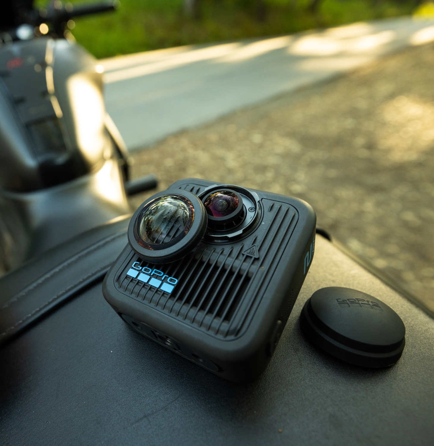 Eine GoPro-Kamera mit abgenommenem Objektiv liegt auf einer schwarzen Oberfläche in der Nähe eines Motorrads. Neben der Kamera liegt eine schwarze Abdeckung.