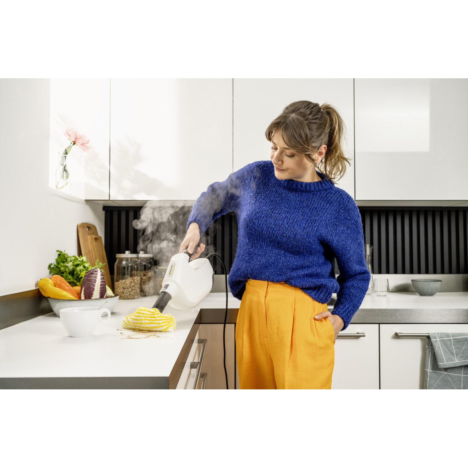Een vrouw maakt een aanrecht schoon met een stoomreiniger. Ze draagt een blauwe trui en een oranje broek. Er liggen groenten in een kom.