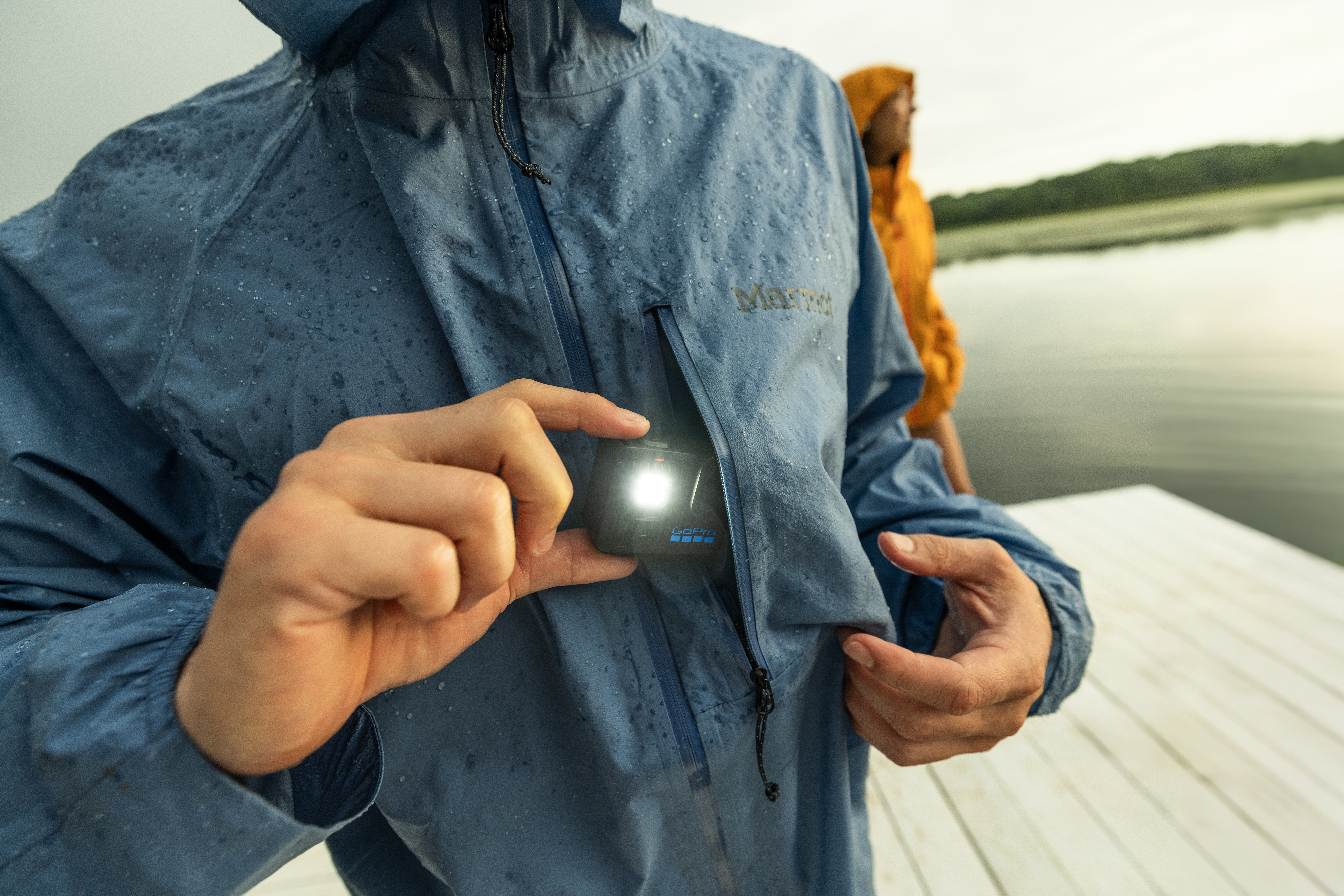 Personne en veste bleue tient une GoPro avec lumière allumée sur un quai. Une autre personne en veste jaune est à l'arrière-plan.