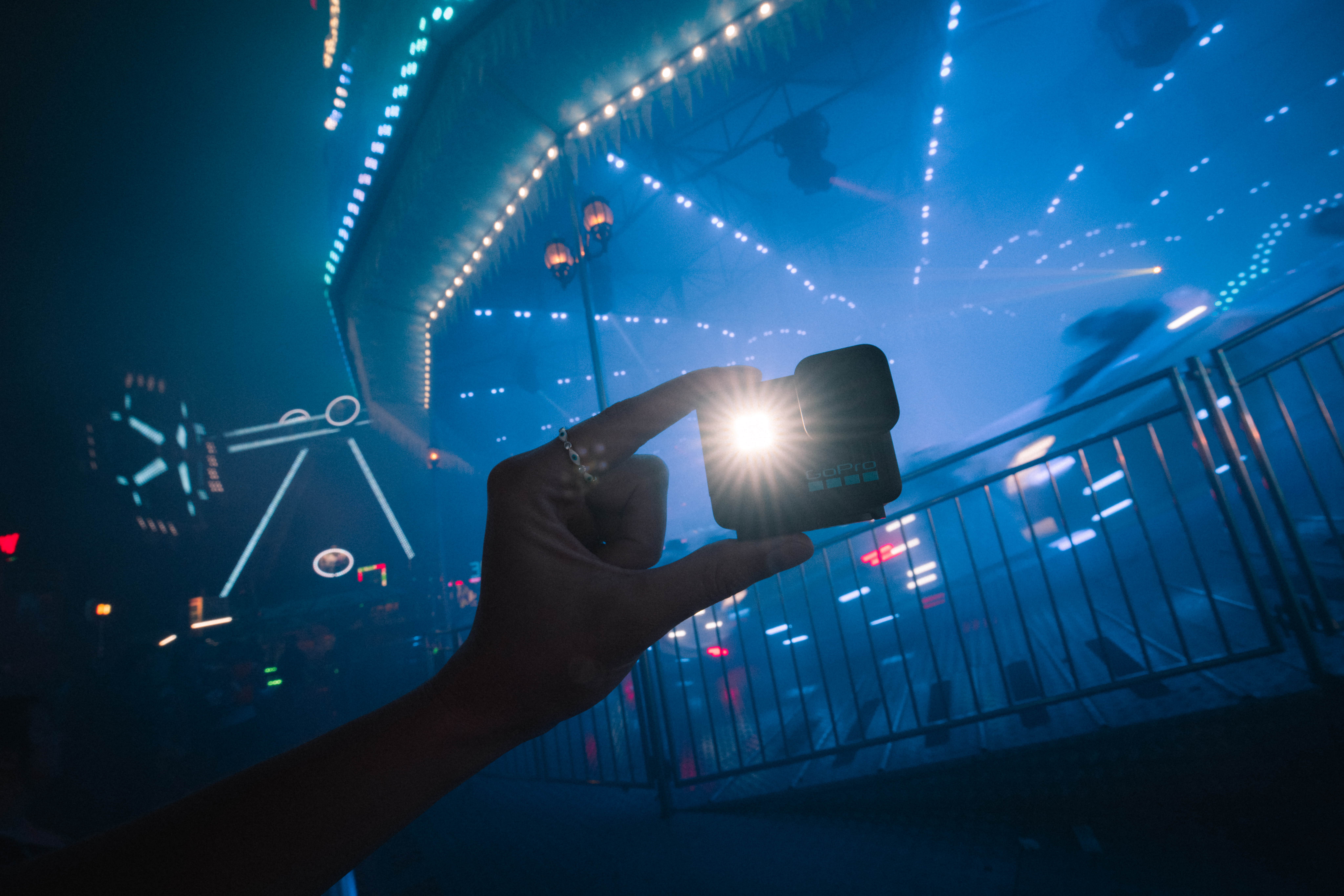 Main tenant une GoPro avec une lumière vive. Grande roue et lumières de parc d'attractions en arrière-plan, créant une atmosphère bleue et sombre.
