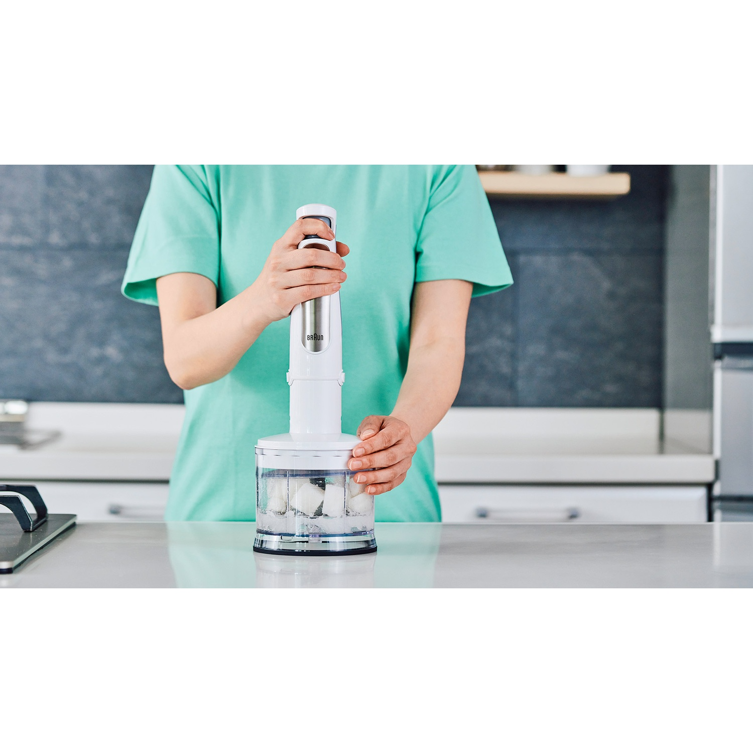 Een persoon in een groen shirt gebruikt een witte blender met ijsblokjes erin. De achtergrond toont een keukenomgeving.