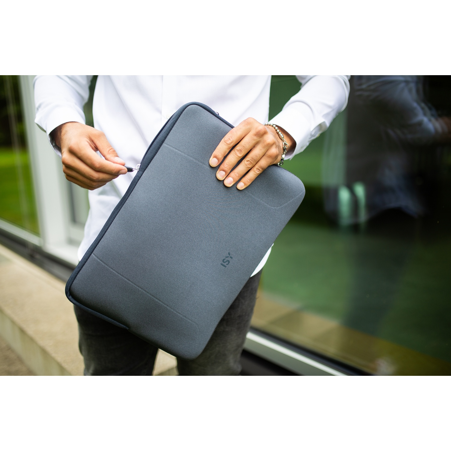 Una persona con una camicia bianca tiene una custodia grigia per laptop. Stanno chiudendo la cerniera. Lo sfondo mostra una scena esterna verde e sfocata.