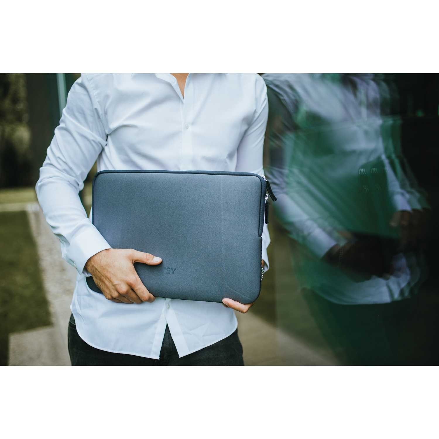 Una persona con camicia bianca tiene una custodia grigia per laptop. Lo sfondo mostra il riflesso della persona e una scena verde all'aperto.