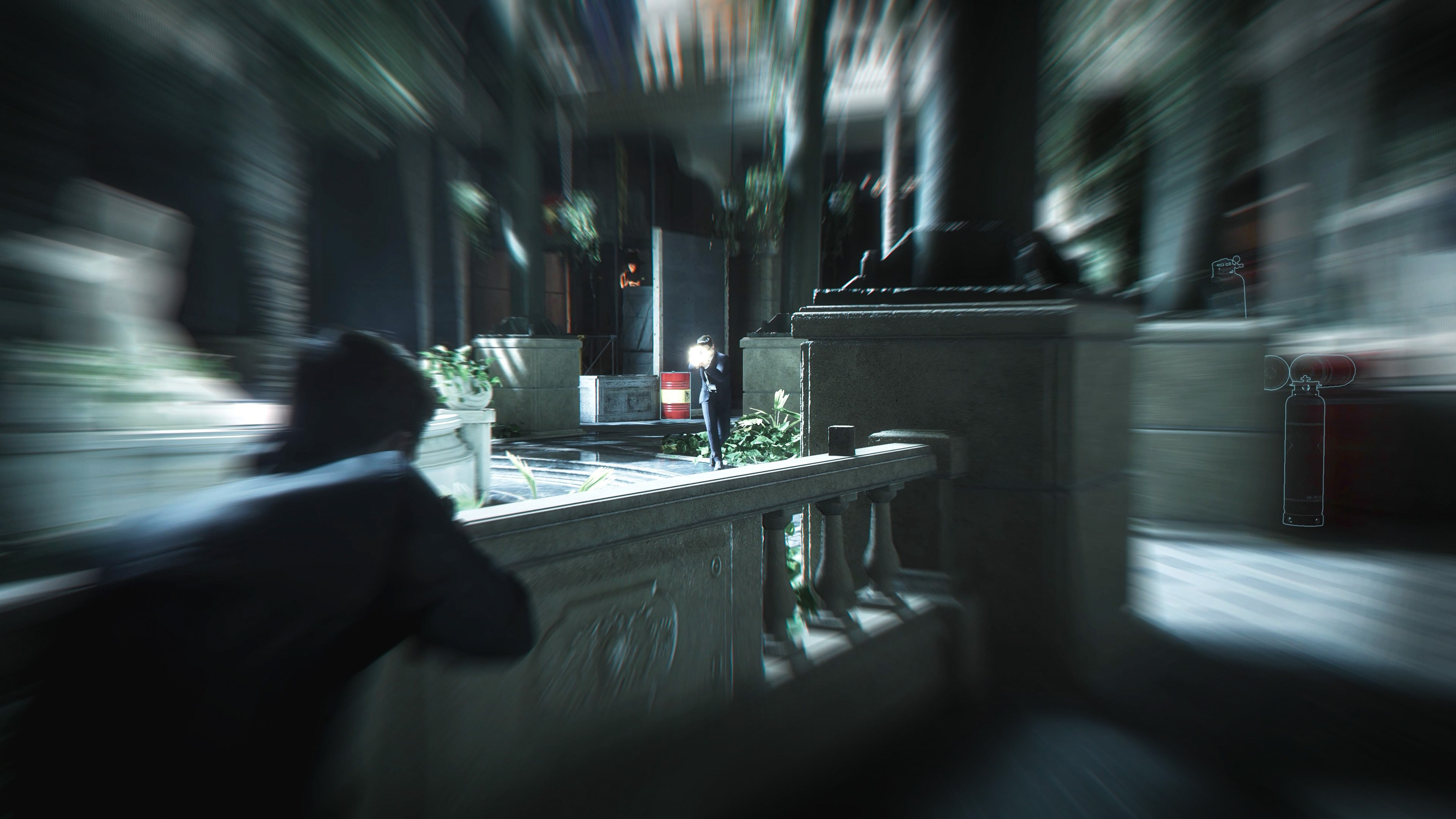 Une personne en costume sombre sur un balcon. Une silhouette floue vise avec une arme. Baril rouge et extincteur en arrière-plan.