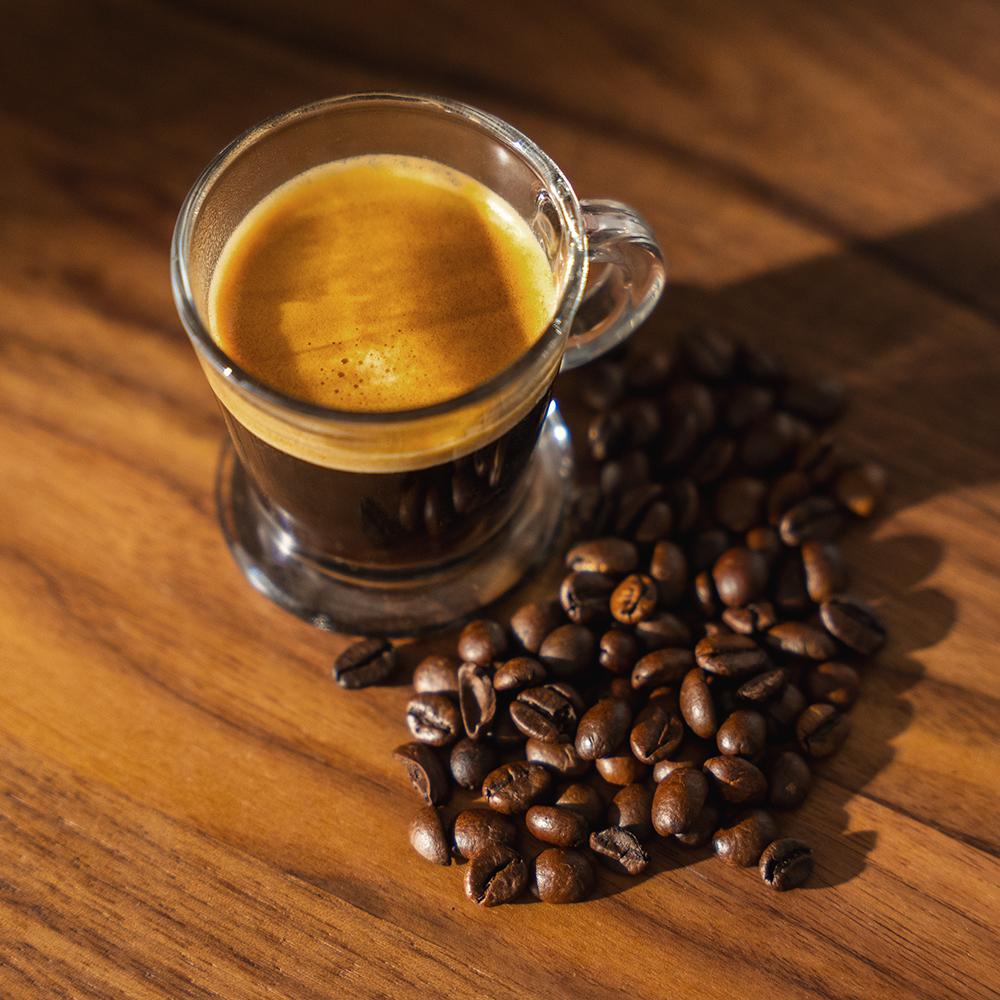 Una taza de espresso con granos de café sobre una mesa de madera. El café tiene una capa de crema. Los granos están esparcidos alrededor.