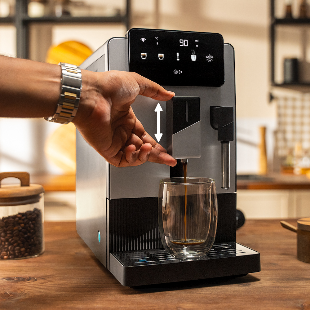 Una persona opera una cafetera plateada. El café se vierte en un vaso. Un tarro de frijoles está a la izquierda.