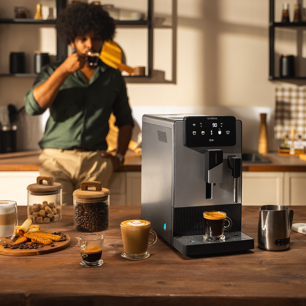 Un hombre bebe café cerca de una cafetera. Tazas de café, galletas, azúcar y granos de café sobre una mesa de madera.