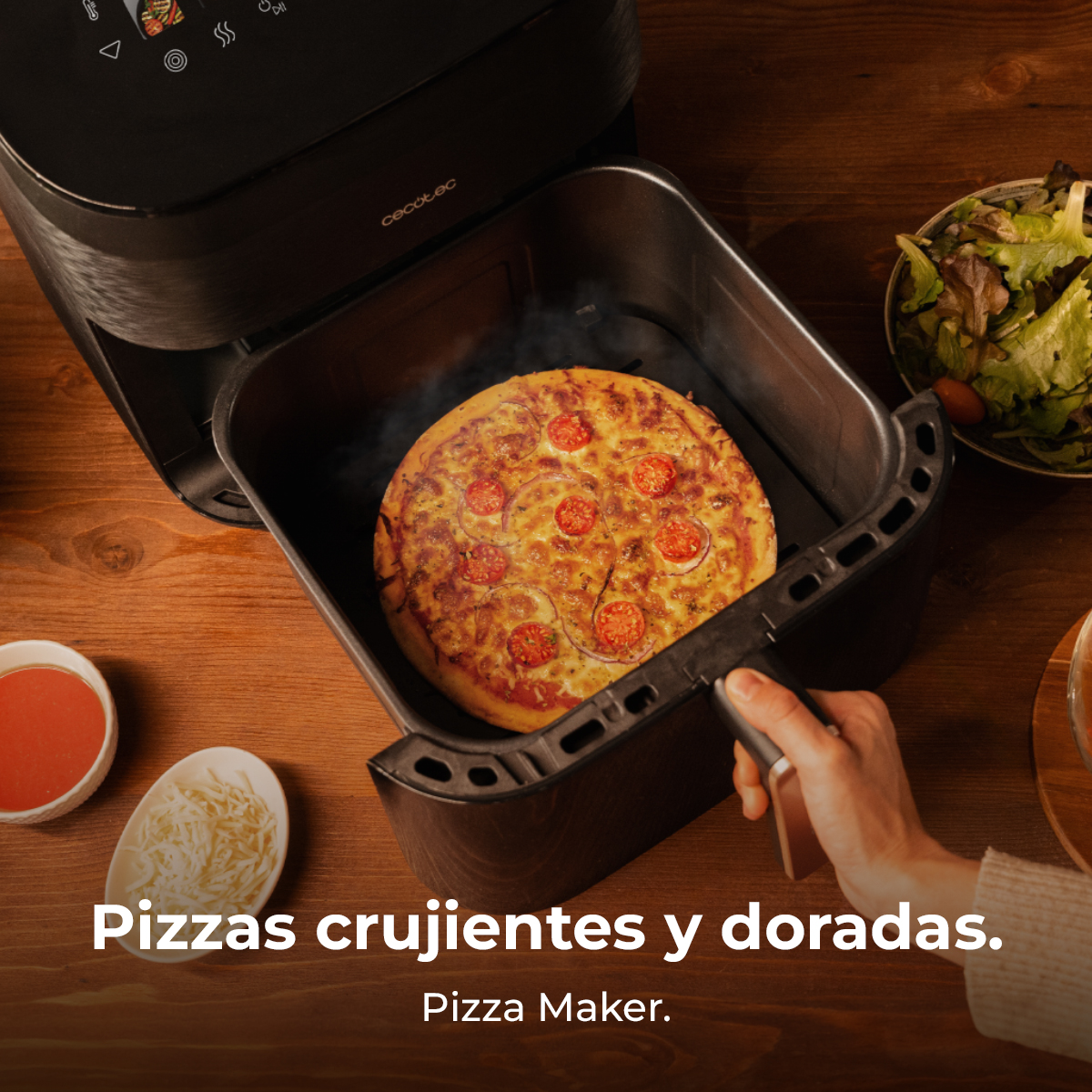 Una pizza cocida con tomates y cebollas está en una freidora de aire. Un bol de ensalada, salsa de tomate y queso están sobre una mesa de madera.