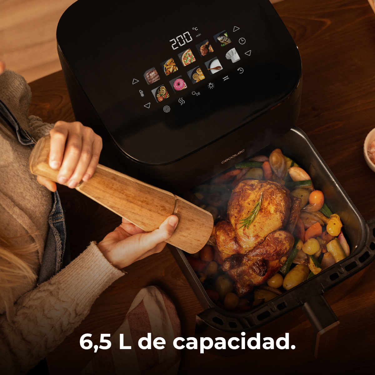 Una persona abre una freidora de aire llena de pollo asado y verduras. La pantalla muestra la temperatura.