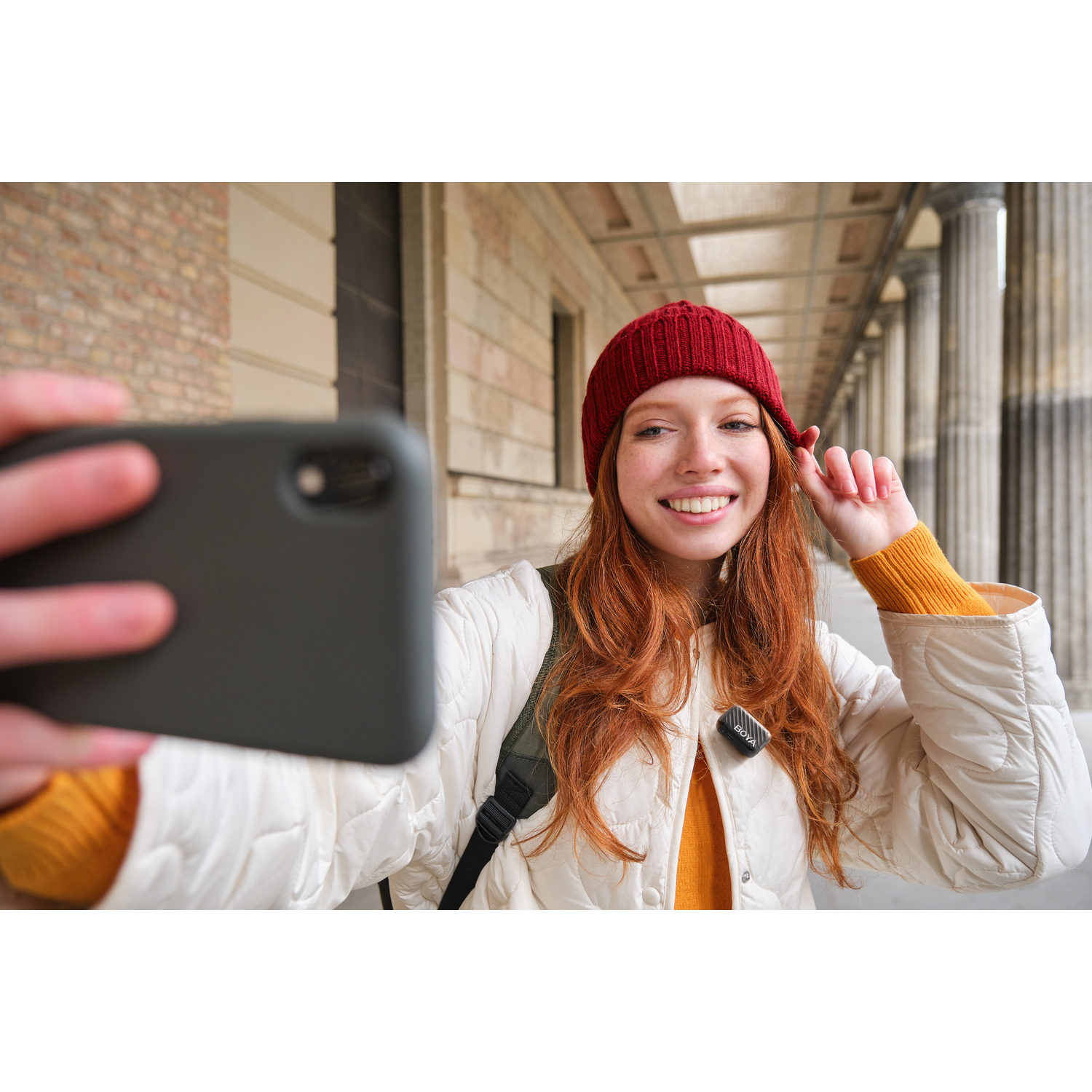 Jonge vrouw met rood haar en een rode hoed maakt een selfie. Ze draagt een witte jas en glimlacht. Achtergrond toont pilaren.