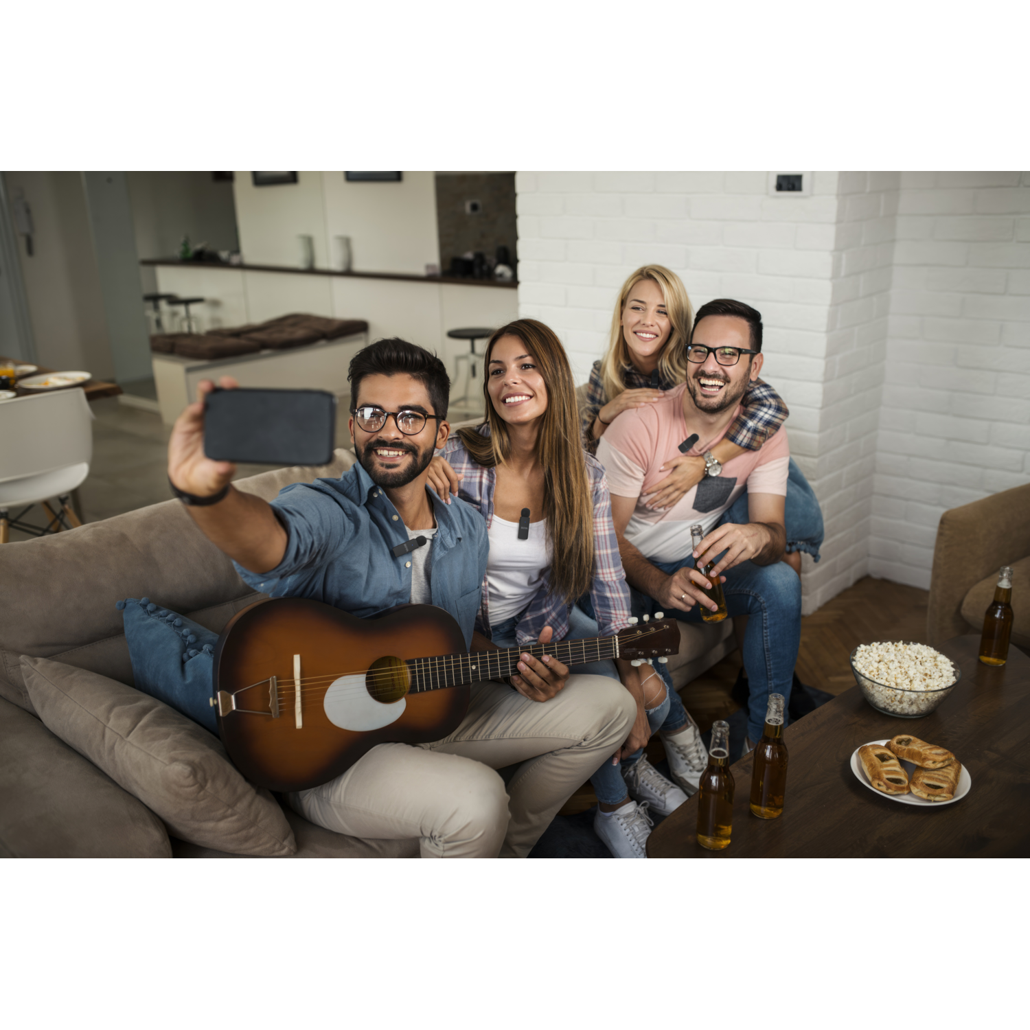 Vier vrienden maken een selfie. Eén houdt een gitaar vast, anderen zitten op een bank met bier en snacks op tafel. Glimlachend.