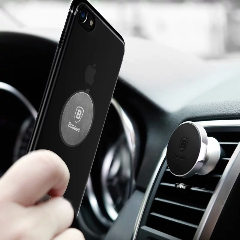 Egy fekete iPhone-t tartanak egy autószellőző tartó közelében. A telefonon egy ezüst Baseus korong van. Egy másik Baseus tartó a szellőzőben van.