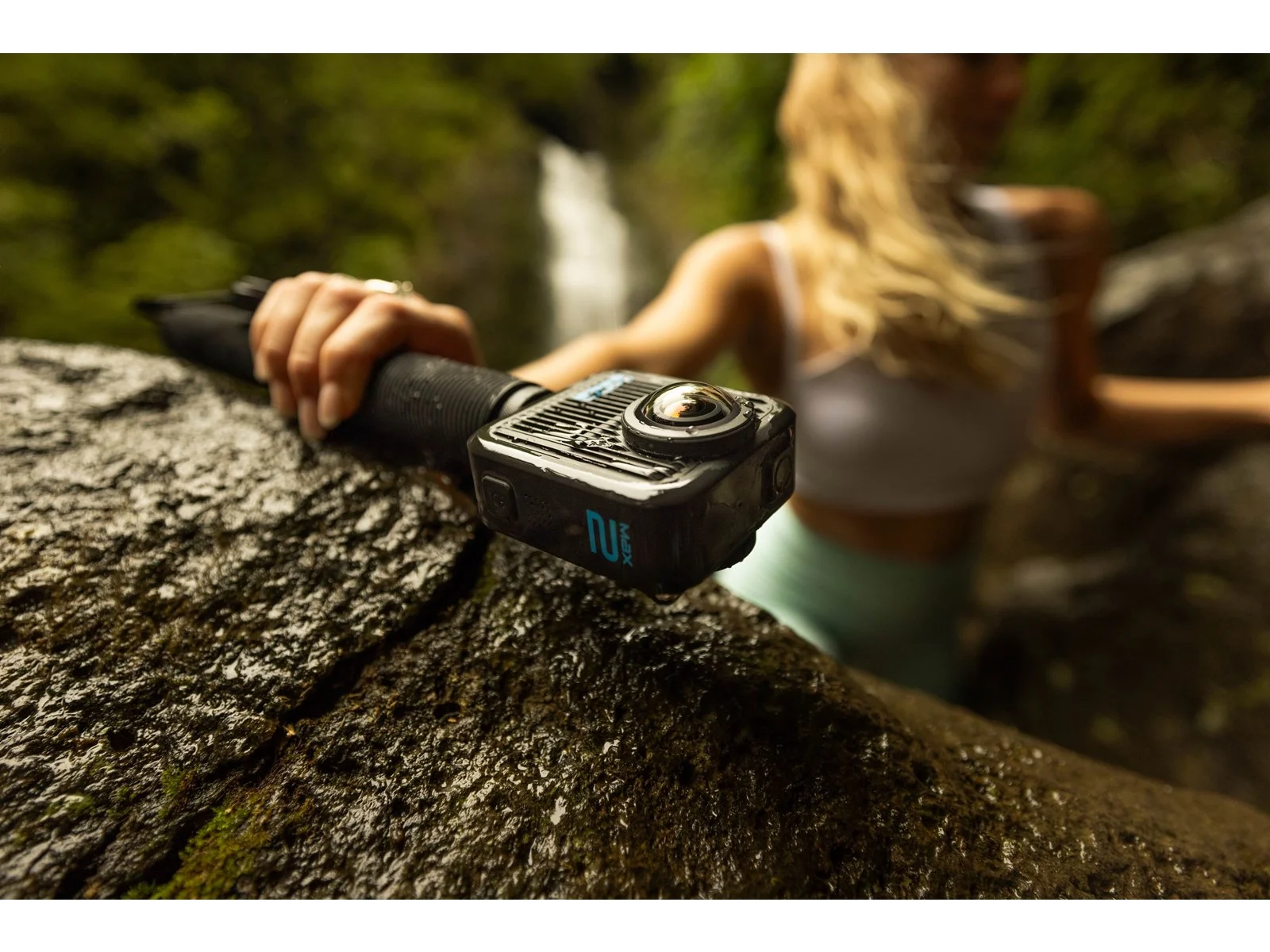 A person holds a black camera on a wet rock. A waterfall is in the background. The person is blurred.