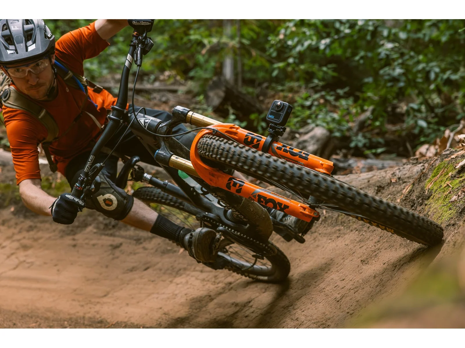 Mountain biker riding a bike on a dirt trail. The bike has orange suspension. The rider wears a helmet and orange shirt.
