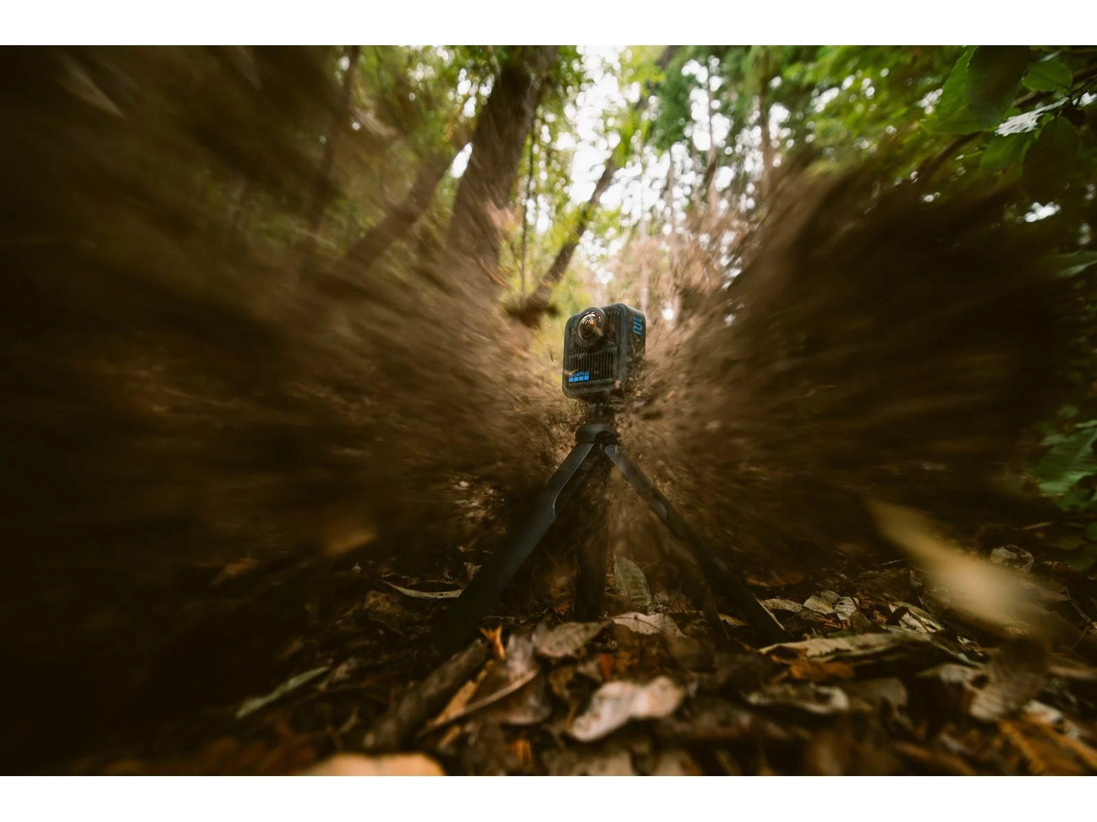 A GoPro camera on a tripod in a forest, surrounded by blurred motion, leaves on ground, trees in background.