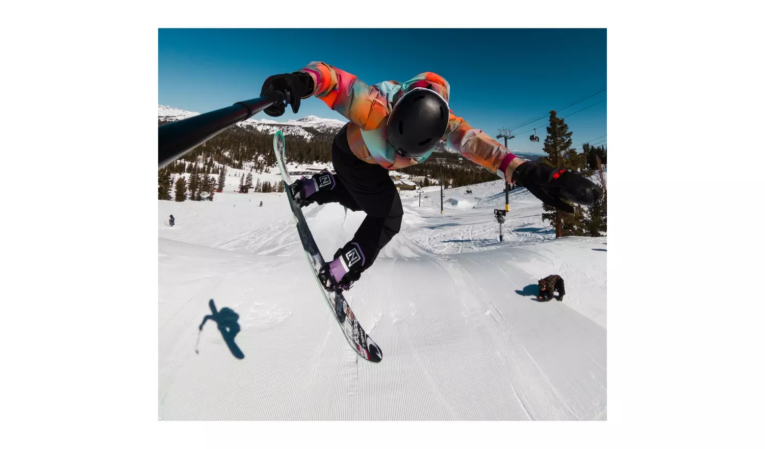 A snowboarder jumps in the air. They wear a colorful jacket, black pants, and helmet. A black dog is in the background.