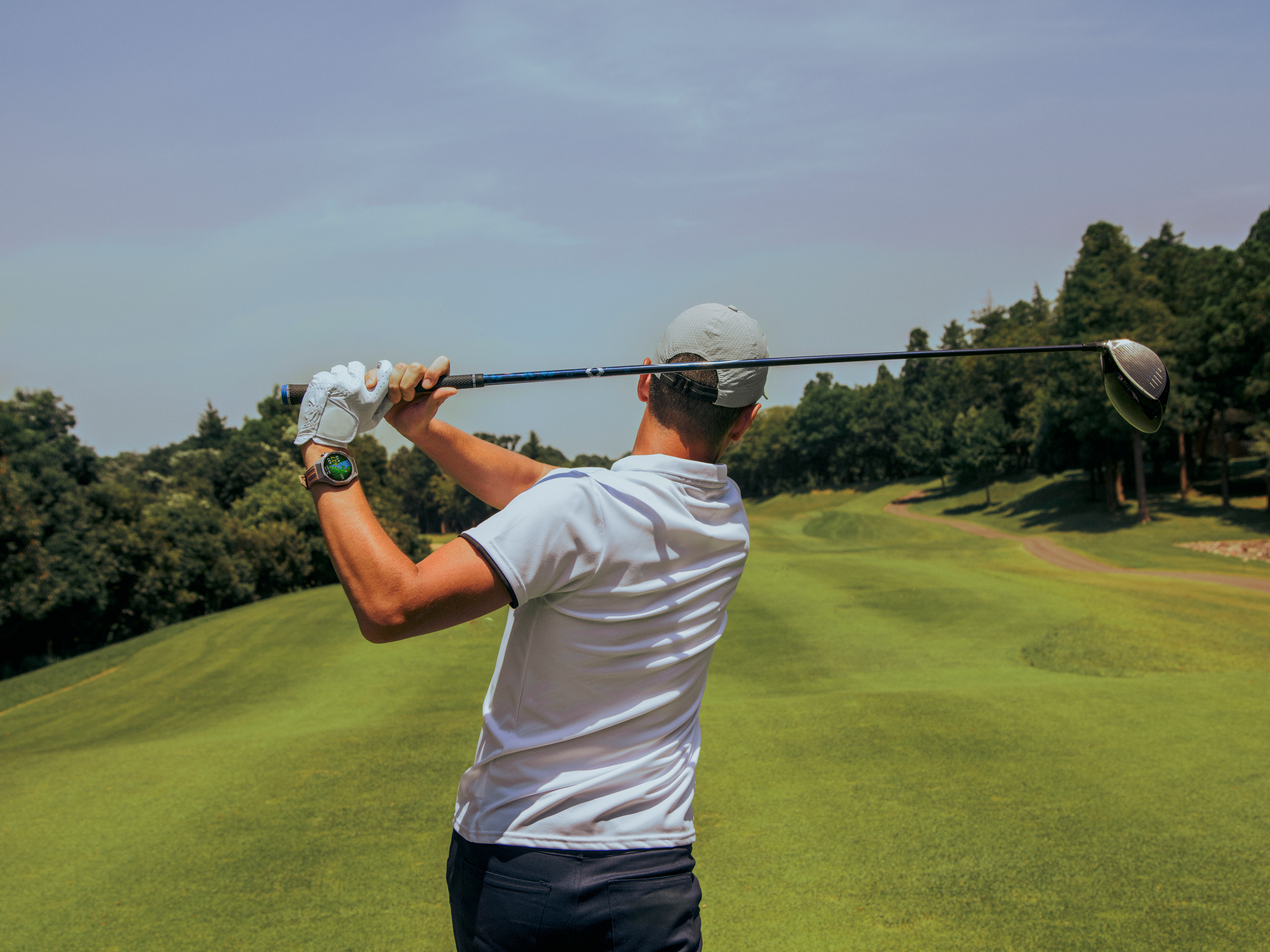 Golfeur balançant un club de golf sur un parcours vert. Arbres et ciel bleu en arrière-plan.