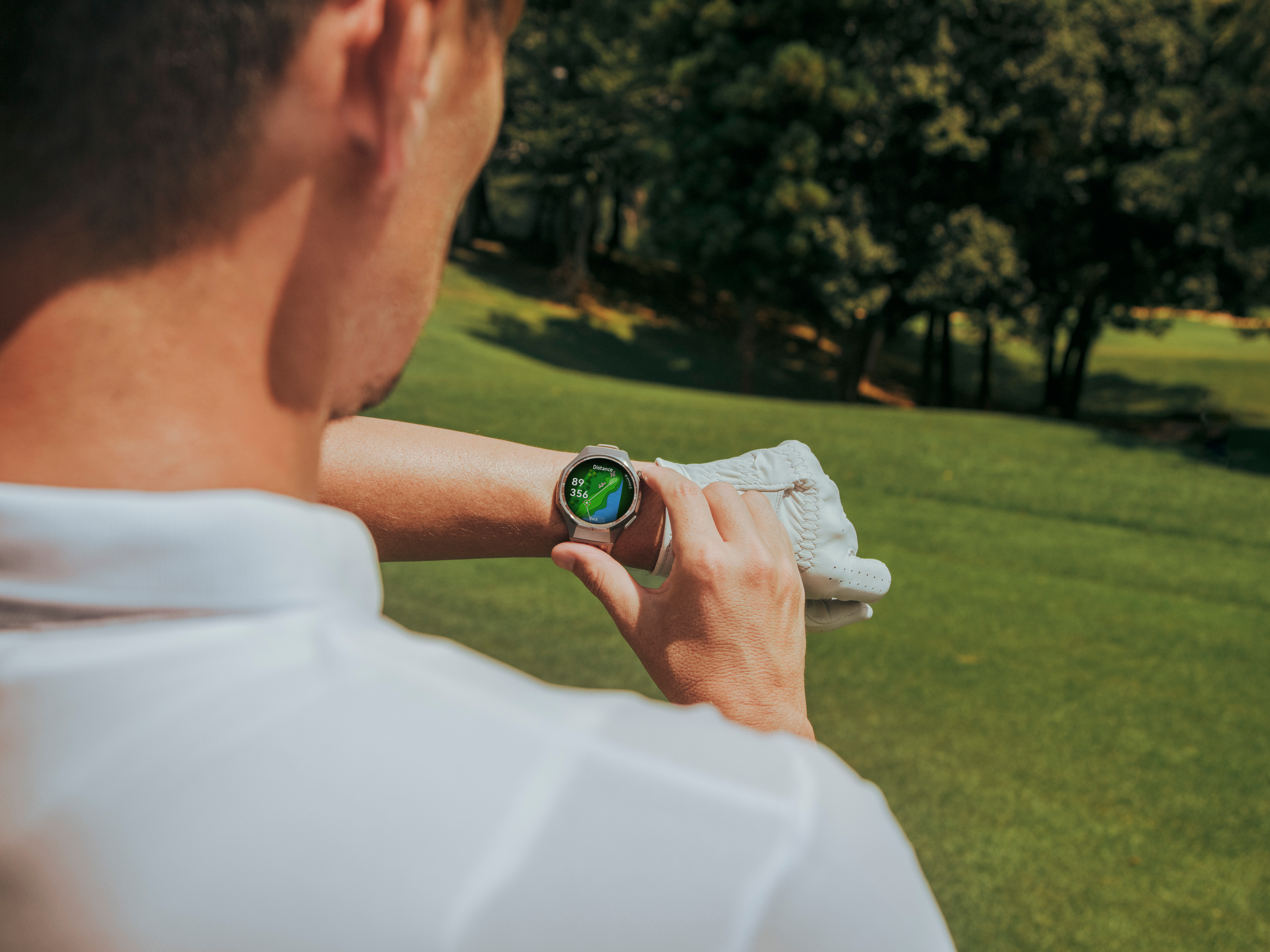 Un homme en chemise blanche consulte une montre intelligente affichant une carte de golf. Il tient un gant de golf. Le fond est de l'herbe verte et des arbres.