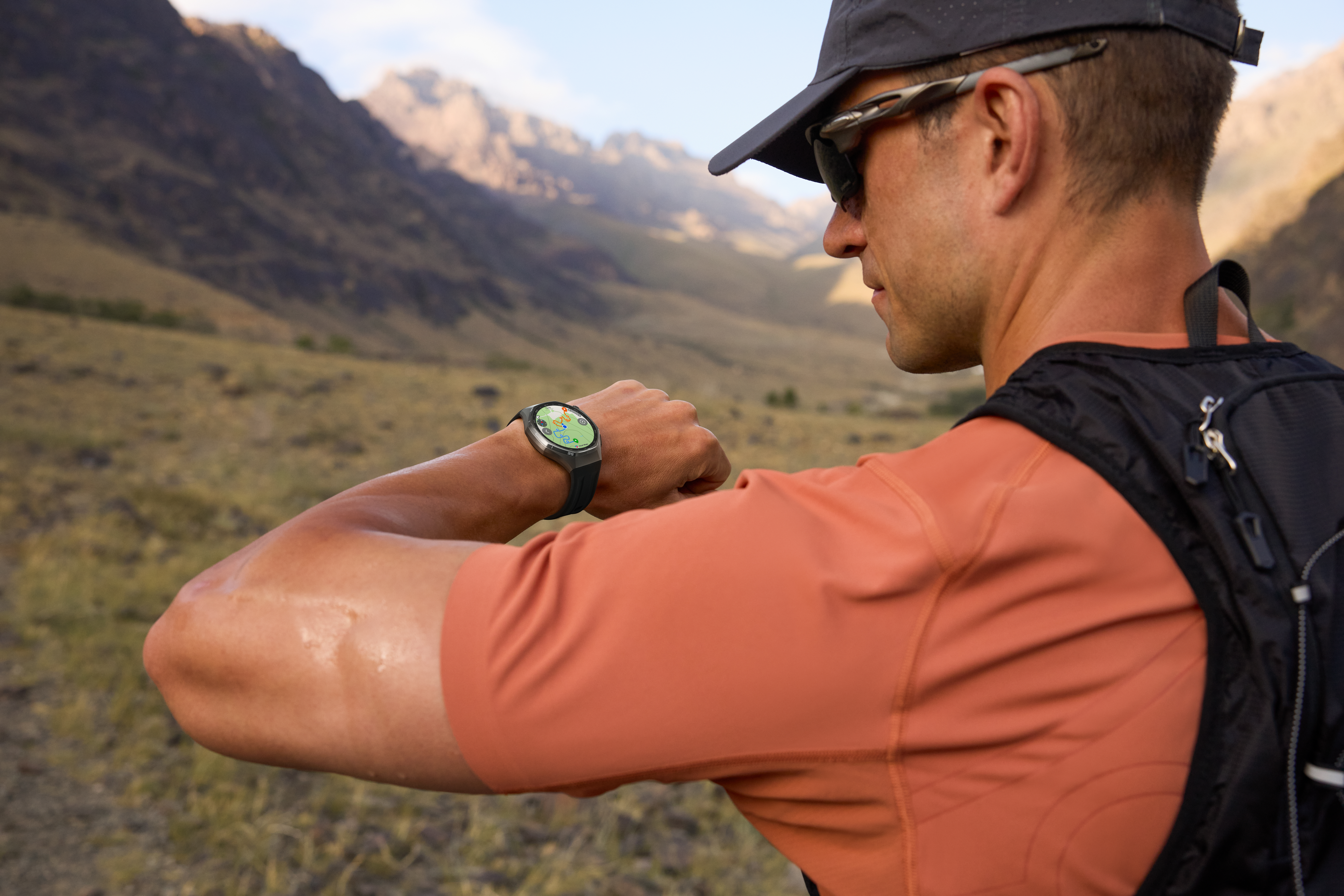 Un homme en casquette et lunettes de soleil consulte sa montre connectée en randonnée. Montagnes et champ en arrière-plan.