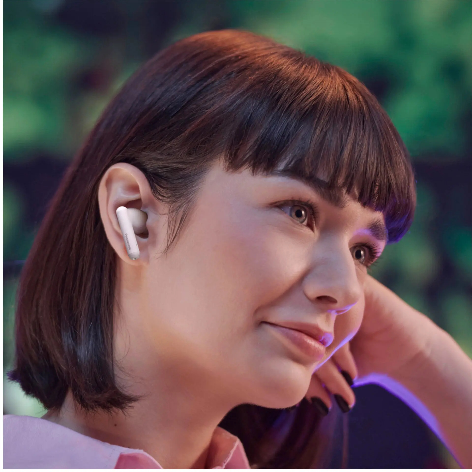 Kobieta ze słuchawkami uśmiecha się, opierając głowę na dłoni. Słuchawka widoczna. Rozmyte zielone i fioletowe tło.