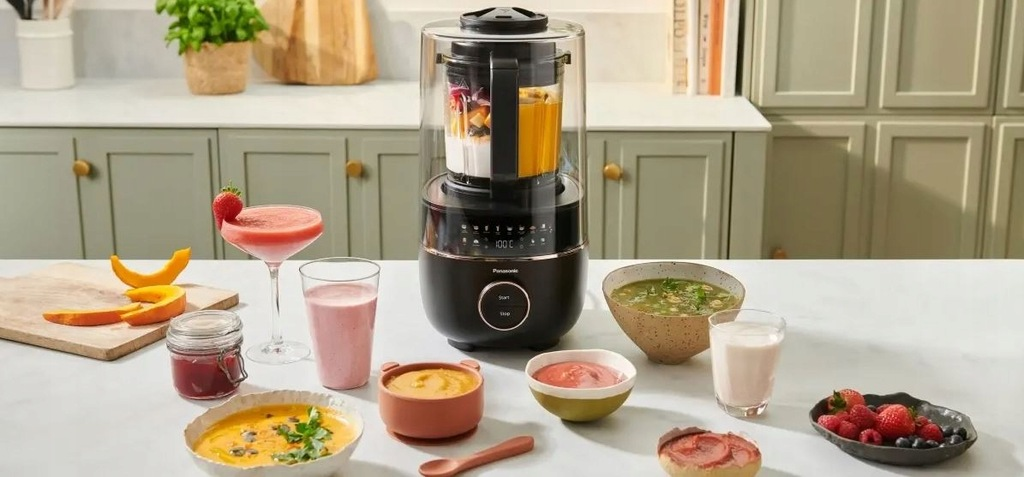 Blender z różnymi przygotowanymi napojami i zupami, plus składniki takie jak owoce i warzywa, ułożone na białej powierzchni.