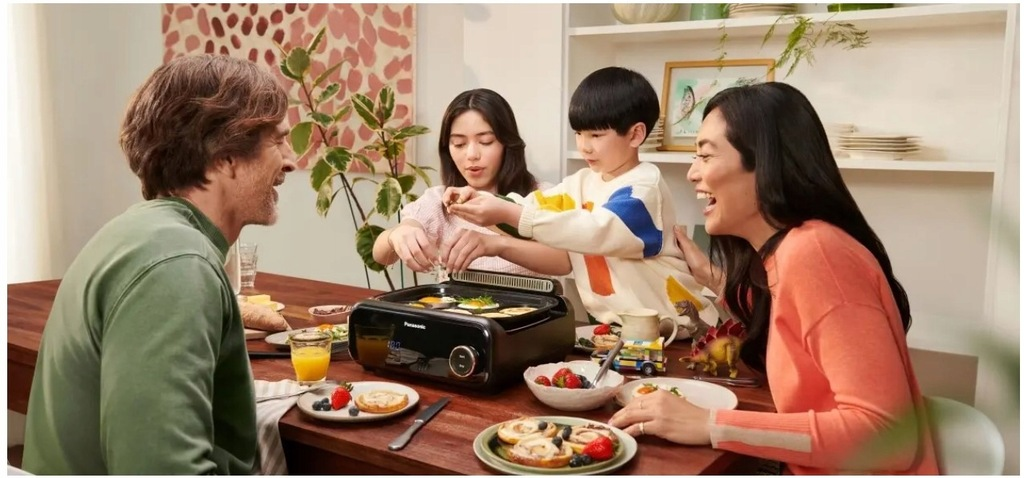 Rodzina przy stole, gotująca jajka na grillu. Uśmiechnięci, z talerzami jedzenia, sokiem i zabawką dinozaurem.