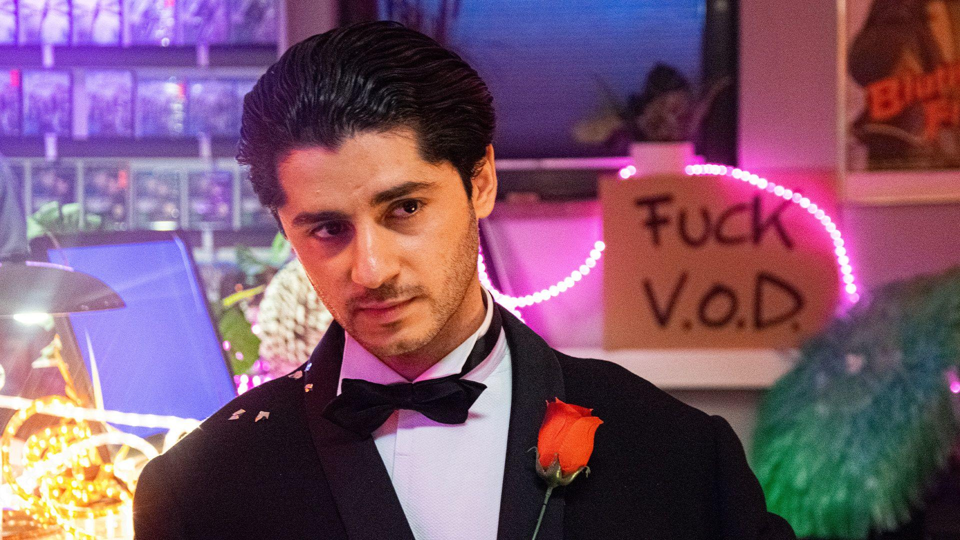 Ein Mann im Anzug mit einer Rose am Revers. Schild mit der Aufschrift 'Fuck V.O.D.'. Neonlichter und Bar-Szene im Hintergrund.