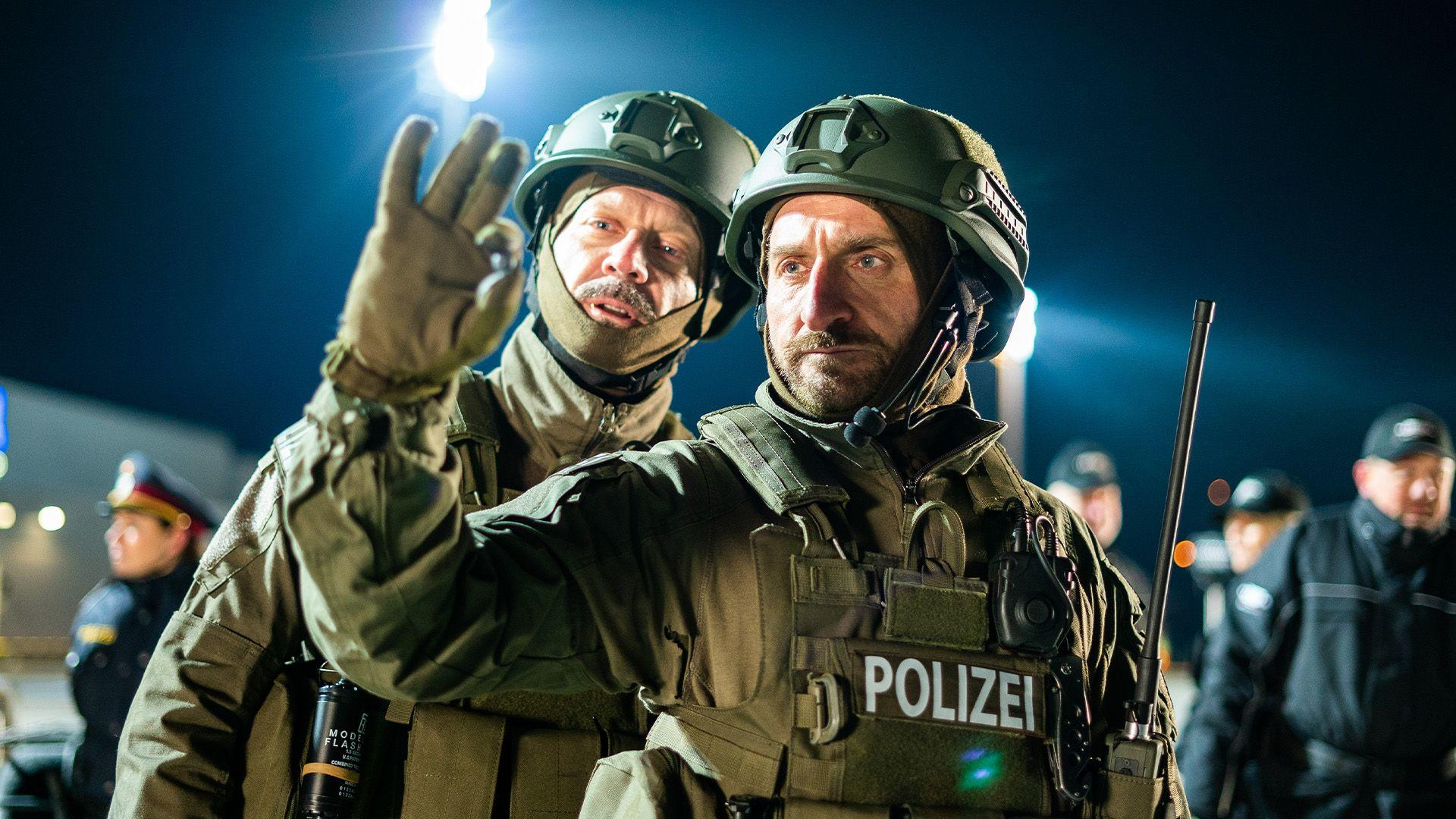 Zwei bewaffnete Polizisten in taktischer Ausrüstung. Einer gestikuliert. POLIZEI ist auf einer Weste zu sehen. Dunkler Hintergrund.