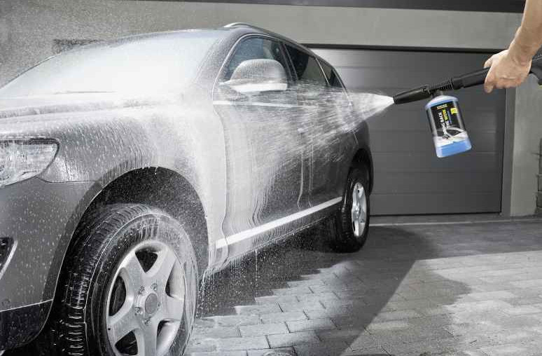 Szary samochód jest myty mydłem. Osoba trzyma narzędzie do mycia. Samochód jest zaparkowany przed garażem.