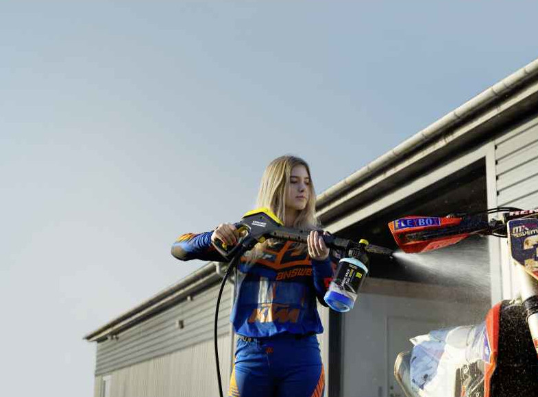 Kobieta w niebiesko-pomarańczowym stroju wyścigowym używa myjki ciśnieniowej do motocykla. Budynek w tle.