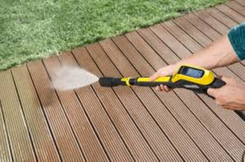 Osoba czyści drewniany taras żółto-czarną myjką ciśnieniową. Woda rozpryskuje się na drewnianych deskach.