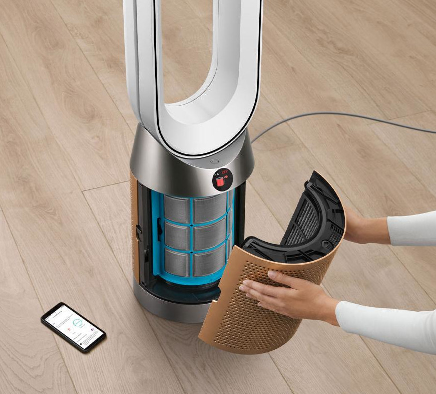 Osoba wymienia filtr oczyszczacza powietrza. Oczyszczacz jest srebrny, brązowy i biały. Na podłodze leży smartfon.