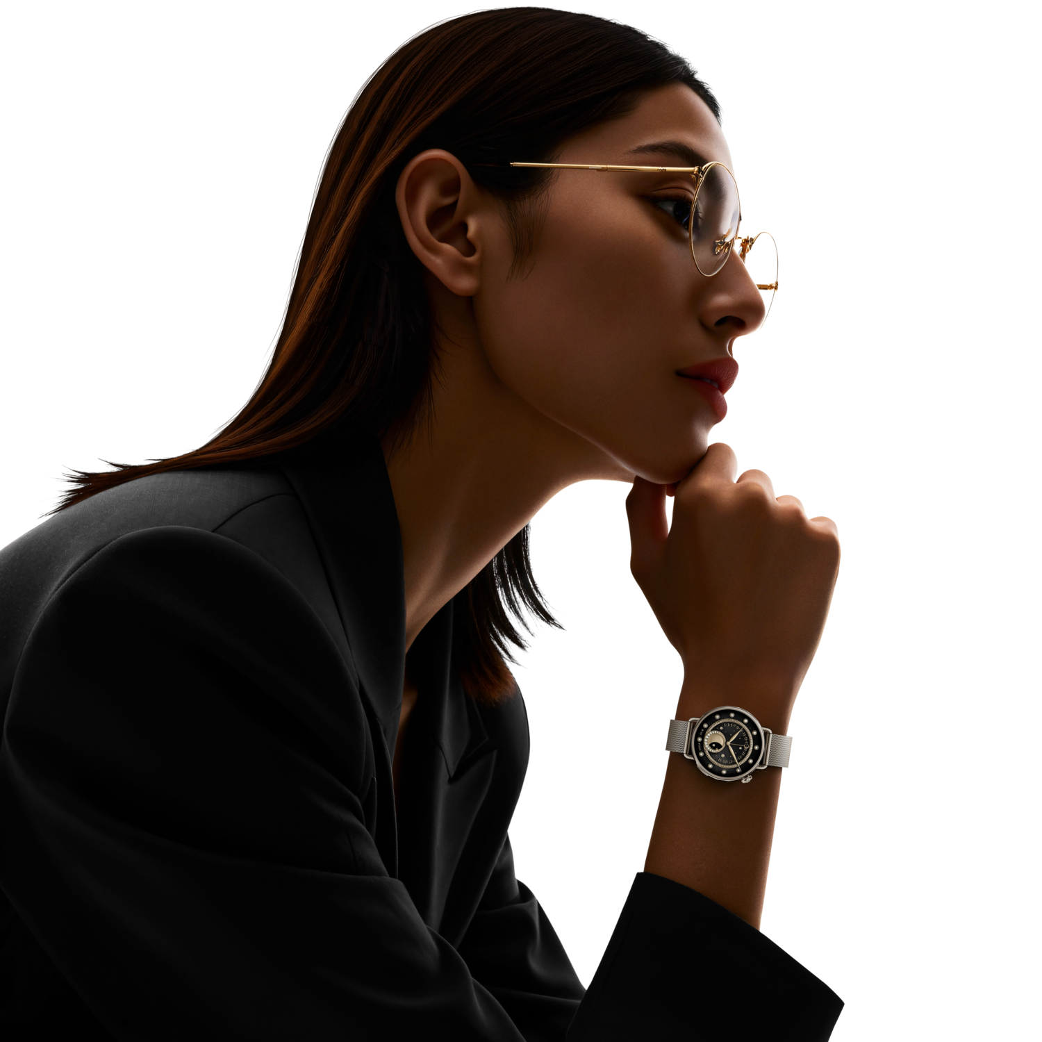 A woman in a black blazer and glasses looks thoughtful. A watch is on her wrist. The background is black.