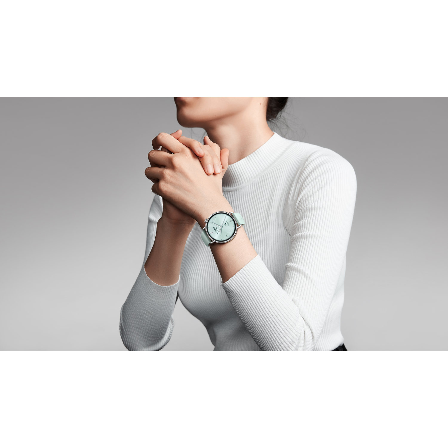 Une femme portant un pull blanc côtelé et une montre vert menthe, mains jointes. Fond gris.