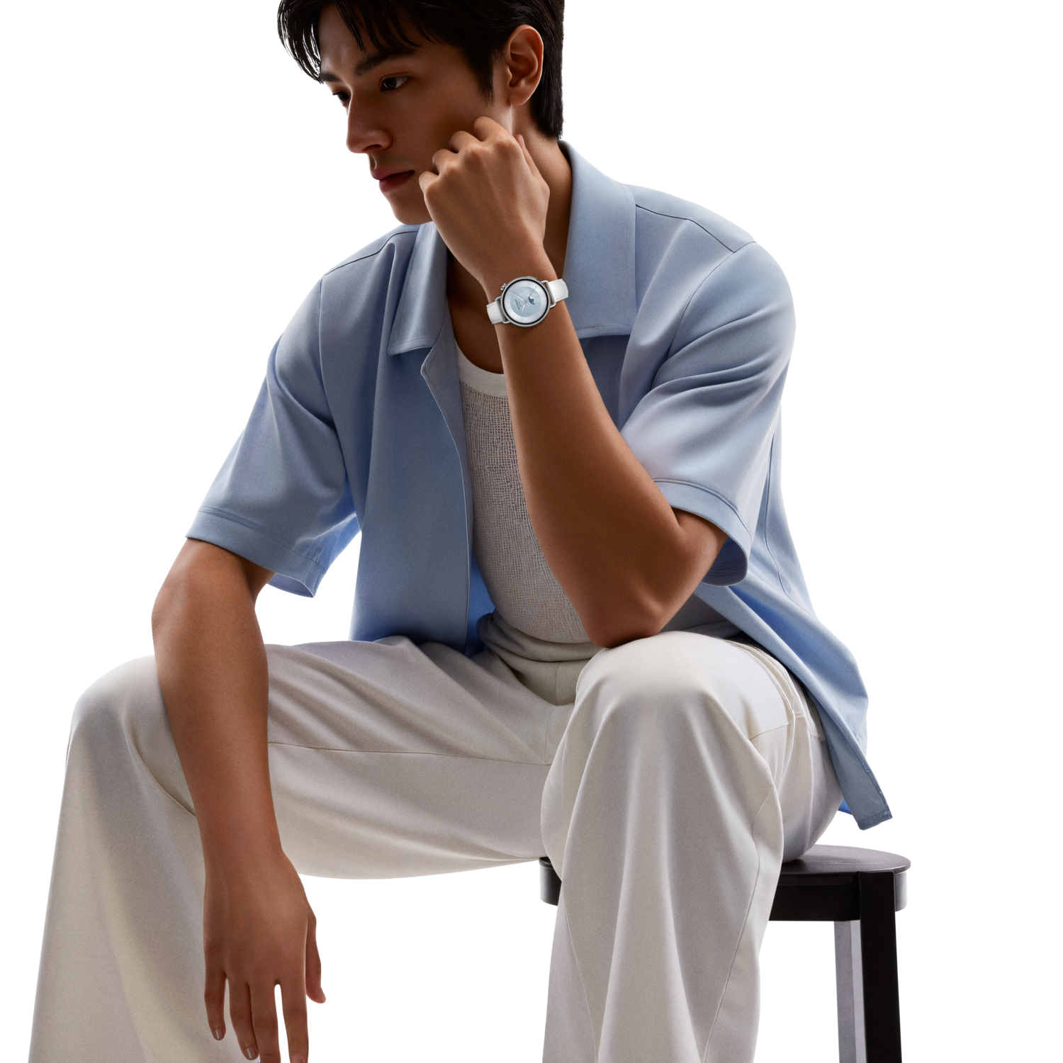A man sits, wearing a watch. He's in light blue shirt and white pants. Black background. He rests his chin on his hand.