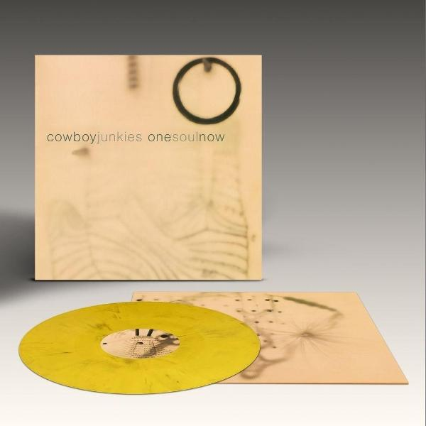 Albumcover mit gelber Vinyl-Schallplatte. Das Cover zeigt einen Ring und den Text 'cowboyjunkies onesoulnow'. Die Platte liegt auf hellbraunem Untergrund.