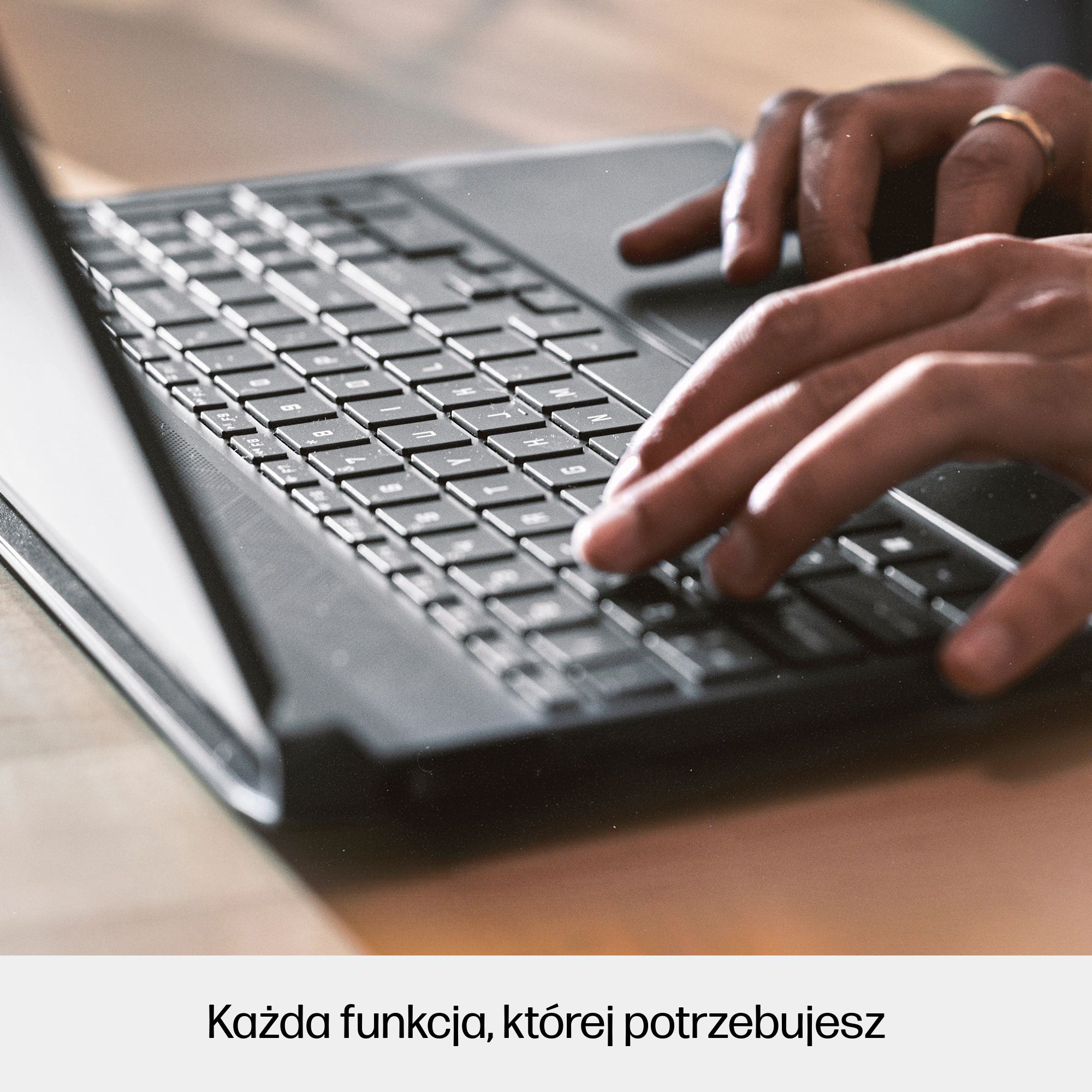 Dłonie piszące na klawiaturze laptopa. Złoty pierścionek na palcu. Laptop leży na drewnianej powierzchni.