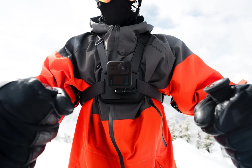 Eine Person in Skiausrüstung mit einer am Brust befestigten Kamera. Sie trägt eine schwarze Maske, eine orangefarbene und graue Jacke und schwarze Handschuhe. Schneeiger Hintergrund.